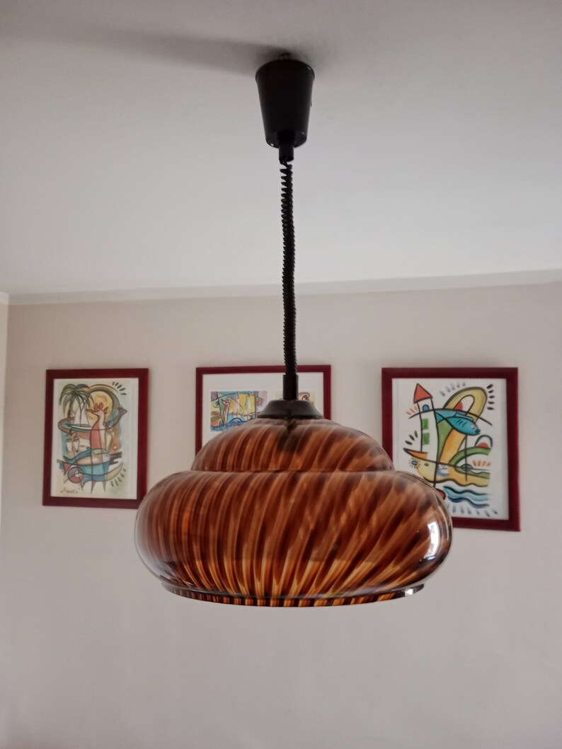 May include: A brown glass pendant light with a swirling pattern hangs from a black cord. The light is suspended from a ceiling in a room with white walls and three framed pictures.