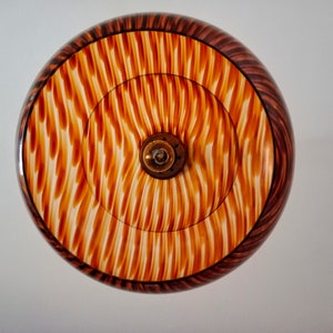 May include: A round, wooden lampshade with a brown and white striped pattern. The lampshade has a central opening for a light bulb.