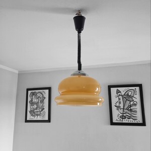 May include: A yellow glass pendant light with a black cord hanging from a white ceiling. Two black framed drawings are on the wall behind the light.