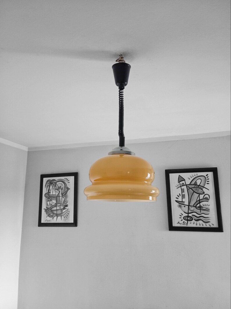May include: A yellow glass pendant light with a black cord hanging from a white ceiling. Two black framed drawings are on the wall behind the light.
