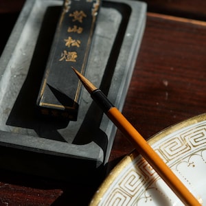 Könnte beinhalten: Ein traditionelles Kalligraphie-Set mit einem rechteckigen Tintenstein, einem schwarzen Tintenstift mit goldener Schrift und einem Kalligraphiepinsel mit Holzgriff. Eine dekorative Platte mit einem goldenen geometrischen Rand ist ebenfalls vorhanden.