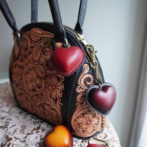 Könnte beinhalten: Eine schwarze und braune Lederhandtasche mit floralem Design und goldenem Reißverschluss. Die Tasche hat zwei schwarze Griffe und ist mit drei herzförmigen Anhängern in Rot und Orange verziert.