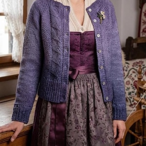 Könnte beinhalten: Eine Frau trägt einen lila Cardigan über einem Kleid. Der Cardigan hat ein Zopfmuster an den Ärmeln und Knöpfe vorne. Das Kleid ist dunkelviolett mit floralem Muster und einem Satinband als Gürtel.