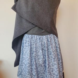 Könnte beinhalten: Ein grauer Umhang mit einer weißen gehäkelten Schleife wird über einem blau-weiß gemusterten Rock mit schwarzer und weißer Spitze getragen.