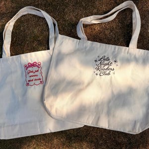 Puede incluir: Dos bolsas de tela color hueso con dos asas. La bolsa izquierda tiene un marco bordado en rosa con el texto "Girls just wanna read books". La bolsa derecha presenta el texto bordado en negro "Late Night Readers Club" con detalles de estrellas.