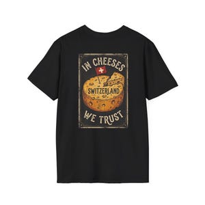May include: A black t-shirt featuring a graphic print of a round Swiss cheese wheel with a small Swiss flag on top. The text reads IN CHEESES SWITZERLAND WE TRUST in a vintage-style rectangular frame, centered on the back of the garment.