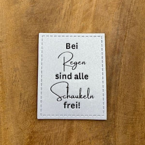 May include: A white sign with black text that reads "Bei Regen sind alle Schaukeln frei!" The sign is on a wooden surface.