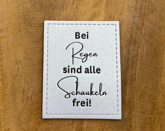 May include: A white sign with black text that reads "Bei Regen sind alle Schaukeln frei!" The sign is on a wooden surface.