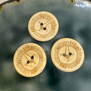 May include: Three wooden buttons with a lion face design. The buttons are light brown with a dark brown lion face and a black nose and eyes.