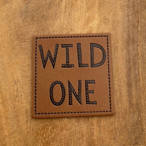 Puede incluir: Un parche de cuero marrón con las palabras "WILD ONE" en letras negras delineadas. Las letras están decoradas con pequeños puntos negros.