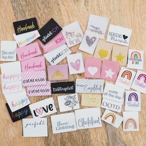 Könnte beinhalten: Eine Sammlung von Stoffetiketten in verschiedenen Farben, darunter Weiß, Rosa und Schwarz. Die Etiketten tragen Texte wie "Handmade", "Happy" und "Love" sowie dekorative Elemente wie Herzen, Sterne und Regenbogen. Die Etiketten liegen auf einer Holzoberfläche.