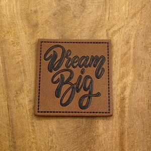 May include: Brown leather patch with stitched edges and the text "Dream Big" in black script.
