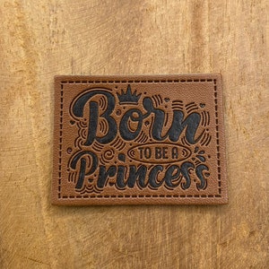 以下が含まれることがあります： 茶色の革のパッチで、黒いステッチが施されています。装飾的なフォントで「Born to be a Princess」というテキストが書かれており、「Born」の文字の上に王冠があります。