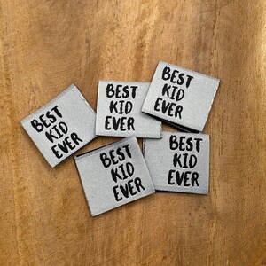 Könnte beinhalten: Sechs graue Stoff-Etiketten mit dem Text "Best Kid Ever" in schwarzer Schrift.