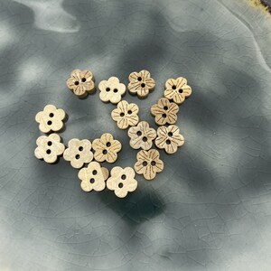 May include: A collection of 14 small wooden buttons shaped like flowers. The buttons are light brown and have a natural wood grain. They are arranged in a loose pile on a blue and white ceramic plate.