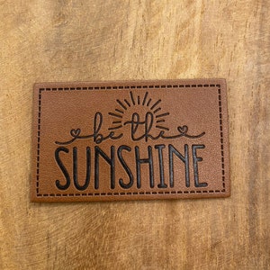 May include: A rectangular, brown leather patch with the phrase "be the SUNSHINE" in black script. The design includes a sunburst above the word "the" and small heart shapes. The patch has a stitched border.