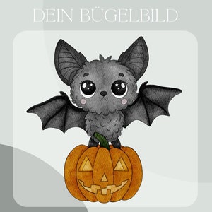 May include: A cute gray bat with big eyes sits on top of an orange pumpkin with a jack-o'-lantern face. The image is titled "Dein Bügelbild" which translates to "Your Iron-on Image".
