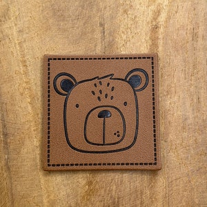 Könnte beinhalten: Ein quadratischer, brauner Lederaufnäher mit einer genähten Umrandung und einem Cartoon-Bärengesicht. Der Bär hat schwarze Augen, Ohren und eine Nase mit kleinen Punkten über den Augen. Der Aufnäher ist wahrscheinlich zum Aufnähen auf Kleidung oder Accessoires gedacht.