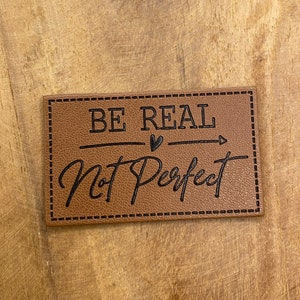 Könnte beinhalten: Braunes Leder-Patch mit dem Text "BE REAL Not Perfect" in schwarzen Buchstaben mit einem kleinen Herzen und einem Pfeil zwischen den Wörtern.