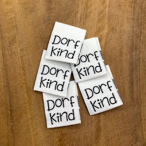 May include: Five white fabric labels with black text that reads "Dorf kind" stacked on top of each other.