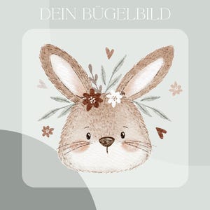 Könnte beinhalten: Ein hellbraunes Hasengesicht mit langen Ohren, verziert mit einem Blumenkranz und Schnurrhaaren. Das Bild enthält den Text "DEIN BÜGELBILD" und "Inklusive Anleitung und Test-Bügelbild" auf hellgrünem Hintergrund. Dekorative Elemente sind kleine Herzen und Blumen.