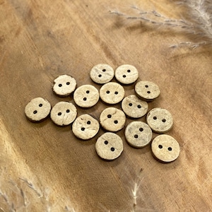 May include: A collection of 15 natural coconut shell buttons in various sizes and shapes. The buttons are a light brown color and have two holes each.