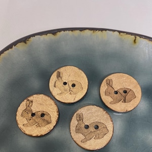 Könnte beinhalten: Vier runde Holzknöpfe mit einem Kaninchen-Design. Die Knöpfe sind hellbraun mit einer lasergeätzten Kaninchensilhouette. Jeder Knopf hat zwei Löcher zum Nähen. Die Knöpfe liegen auf einem blauen Keramikteller.