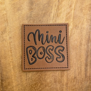Pode incluir: Um remendo de couro marrom com o texto "mini boss" em letras pretas. O remendo tem uma borda costurada.