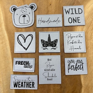 Può includere: Dieci toppe da stirare con diversi disegni e motti. Le toppe sono argentate con testo e disegni neri. I disegni includono un orso, un cuore, un unicorno, un bassotto e le parole "Wild One", "Handmade", "You are perfect", "Sweater Weather", "Frech Dachs", e "Bei Regen sind alle Schaukeln frei!"