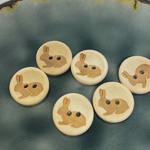 Puede incluir: Seis botones de madera con fondo beige claro y diseño de conejo marrón. Cada botón tiene dos pequeños agujeros para coser. Los botones están colocados sobre un plato de cerámica turquesa.