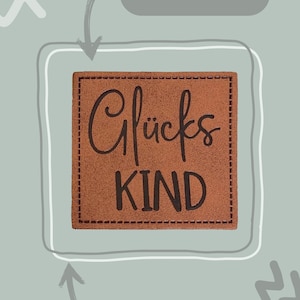 Könnte beinhalten: Ein braunes Leder-Etikett mit dem Text "Glücks KIND" in schwarzen Buchstaben. Das Etikett ist 4 cm x 4 cm groß und hat Nähte an den Rändern. Es ist zum Annähen an Stoff gedacht.
