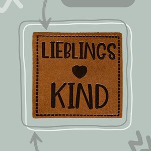 Könnte beinhalten: Braunes Lederlabel mit dem Text "LIEBLINGS KIND" und einem Herzsymbol. Das Label ist 4 cm x 4 cm groß und hat Nähte an den Rändern.