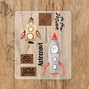Puede incluir: Un conjunto de cuatro pegatinas con cohetes y texto con temática espacial. Las pegatinas están sobre un fondo de madera. El texto en las pegatinas dice "Reach for the stars", "Little Explorer", "Astronaut" y "To the moon".