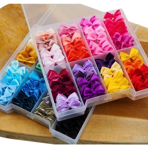 May include: A clear plastic storage box filled with a variety of colorful satin bows. The bows are arranged in rows and columns, with each color separated into its own compartment. The box is sitting on a wooden surface.
