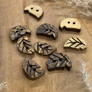 May include: A collection of leaf-shaped buttons in various stages of completion. The buttons are made of wood and feature a natural, light brown color with darker brown accents. Each button has two holes for sewing.