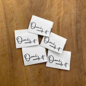 Könnte beinhalten: Fünf weiße Stoff-Etiketten mit schwarzem Text, der "Omi made it" lautet.