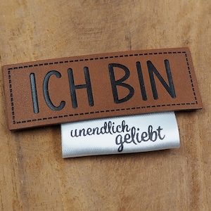 May include: Brown leather patch with black stitching and the text "ICH BIN" in black lettering. A white satin label with black text that reads "unendlich geliebt" is attached to the leather patch.
