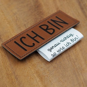 Könnte beinhalten: Ein braunes Leder-Patch mit dem Text "ICH BIN" in schwarzen Buchstaben. Ein weißes Stoff-Etikett mit dem Text "genau richtig, so wie ich bin!" ist unter das Leder-Patch gesteckt.