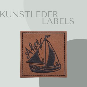 Könnte beinhalten: Ein quadratisches, braunes Lederetikett mit einer schwarzen Segelboot-Illustration und dem Wort "Ahoi" in Schreibschrift. Das Etikett ist an den Rändern genäht und trägt oben den Text "KUNSTLEDER LABELS".