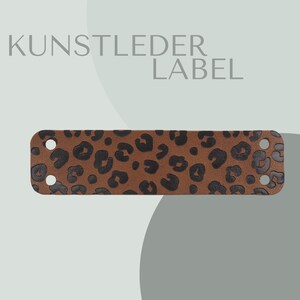 Könnte beinhalten: Ein braunes Lederetikett mit einem schwarzen Leopardenmuster. Das Etikett hat abgerundete Ecken und zwei Löcher an jedem Ende. Der Text "KUNSTLEDER LABEL" ist über dem Etikett aufgedruckt.