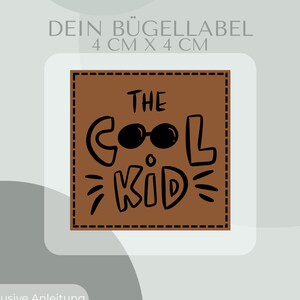 Peut inclure: Un écusson carré marron avec le texte "THE COOL KID" en noir, avec des lunettes de soleil sur le "O". L'écusson mesure 4 cm x 4 cm. Le texte supplémentaire comprend "DEIN BÜGELLABEL" et "Inklusive Anleitung und Test-Bügelbild".