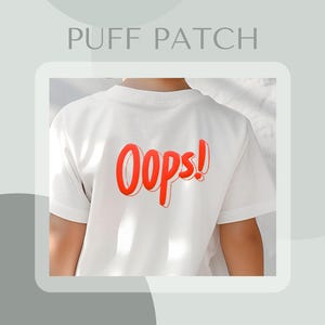 Pode incluir: Camiseta branca com um gráfico vermelho "Oops!" nas costas. O gráfico está em um estilo volumoso, 3D.