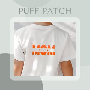 Könnte beinhalten: Weißes T-Shirt mit einem orangenen "MOM"-Design mit zwei kleinen Herzen über dem "O". Der Text ist in einer fetten, blockartigen Schrift.
