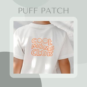 Könnte beinhalten: Weißes T-Shirt mit einer orangefarbenen Grafik, auf der "COOL MOMS CLUB" mit Smileys steht.