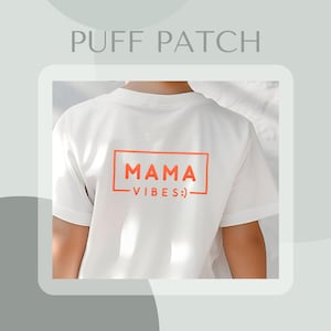 Puede incluir: Camiseta blanca con un parche naranja "MAMA VIBES :)" en la espalda.