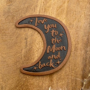 Könnte beinhalten: Ein braunes Leder-Halbmond-Patch mit dem Text "Love you to the moon and back" in schwarzer Schrift. Das Patch hat kleine Stern-Akzente.