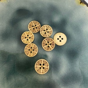 May include: A collection of six wooden buttons with a decorative, intricate design. The buttons are light brown with a dark brown design and four holes each.