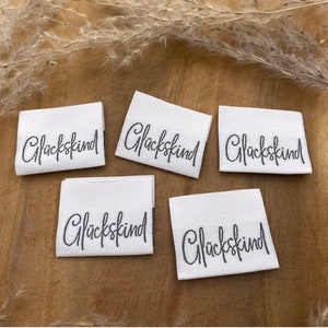 Könnte beinhalten: Fünf weiße Stoff-Etiketten mit dem Wort "Glückskind" in schwarzer Schrift.