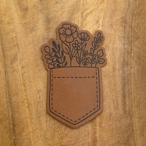 Könnte beinhalten: Ein braunes Lederstück mit einem schwarzen Umriss einer Tasche und Blumen. Die Tasche ist mit einer gestrichelten Linie genäht.