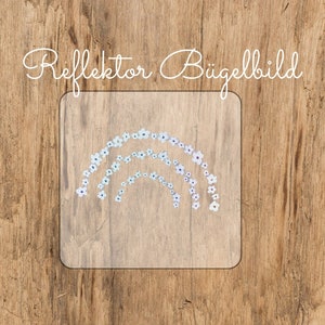 Pode incluir: Uma folha de transferência termocolante transparente com um design de arco-íris feito de flores brancas. O texto "Reflektor Bügelbild" está impresso acima do design.